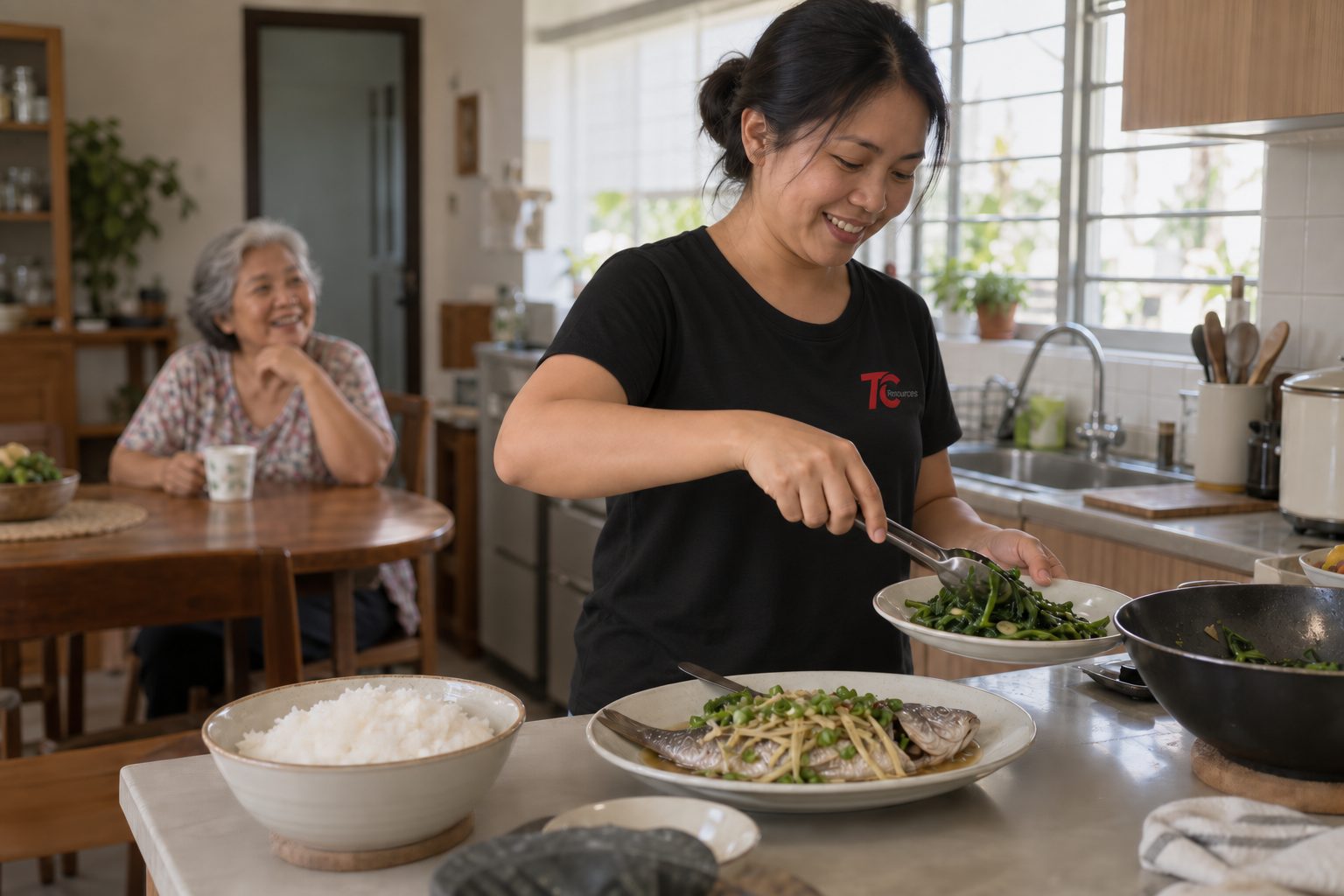 A helper preparing a meal in a Malaysian kitchen while an elderly woman sits at the table behind her